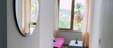 Un couloir étroit, avec un grand miroir sur le mur de gauche, un lit recouvert d’une couverture rose et une petite table sur laquelle est posé un vase, au centre.