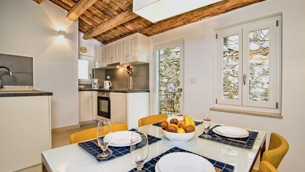 A dining area with a white table set for four, a bowl of fruit, and a view of a stone wall outside. - Stunning old town apartment with private terrace, outdoor shower - only 500 meters to the beach (Rovinj)