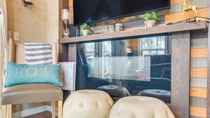 Un salon chaleureux avec un mur en bois, un téléviseur à écran plat fixé au-dessus d’une cheminée, et un coin salon confortable avec un fauteuil et des poufs.