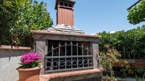 Un horno exterior de piedra con una parrilla metálica, rodeado de follaje y una maceta con flores rosas.