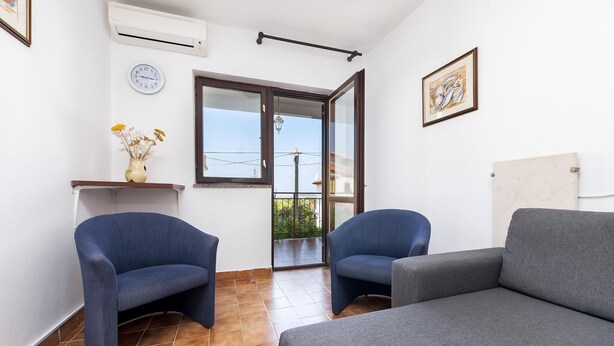Une pièce avec deux fauteuils bleus, un canapé gris, une horloge murale, un tableau encadré et une fenêtre donnant sur un paysage verdoyant.