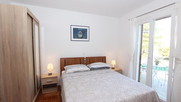 Un dormitorio con cama, dos mesitas de noche con lámparas, un armario de madera, un cuadro enmarcado en la pared y un ventanal corredero que da a un espacio exterior.
