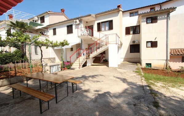 A two-story building with a staircase leading to a balcony, a concrete patio with a wooden table, and a garden area with plants and a pergola.