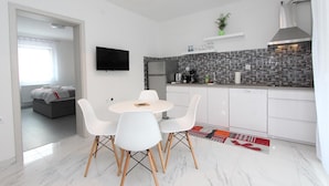 Une cuisine moderne avec des meubles blancs, un pare-feu en carrelage et un four encastré. On y trouve un coin repas avec une table et des chaises blanches, ainsi qu’un petit îlot de cuisine.