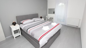 Une chambre à coucher moderne avec un lit gris, des tables de chevet blanches, un bureau et une grande fenêtre ornée de rideaux légers.