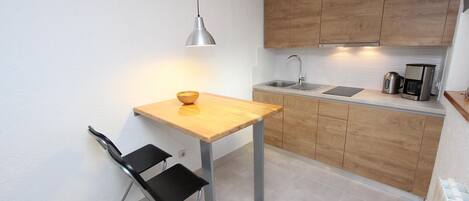 A compact kitchen with a wooden table, a black chair, a hanging light, and a coffee maker.