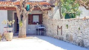 Um edifício de paredes de pedra com telhado de telhas, uma árvore em primeiro plano e uma mesa com cadeiras na parte externa.