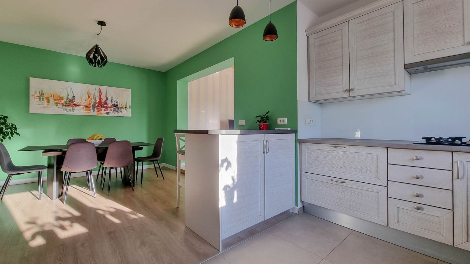 Eine moderne Küche mit weißen Schränken, eine Essecke mit Tisch und Stühlen sowie eine grüne Wand mit einem Gemälde.
