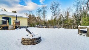 A snow-covered yard with a wooden deck, a stone fire pit, and a house with a green door. - Near Skiing: Serene Harrison Retreat w/ Fire Pit (Harrison)