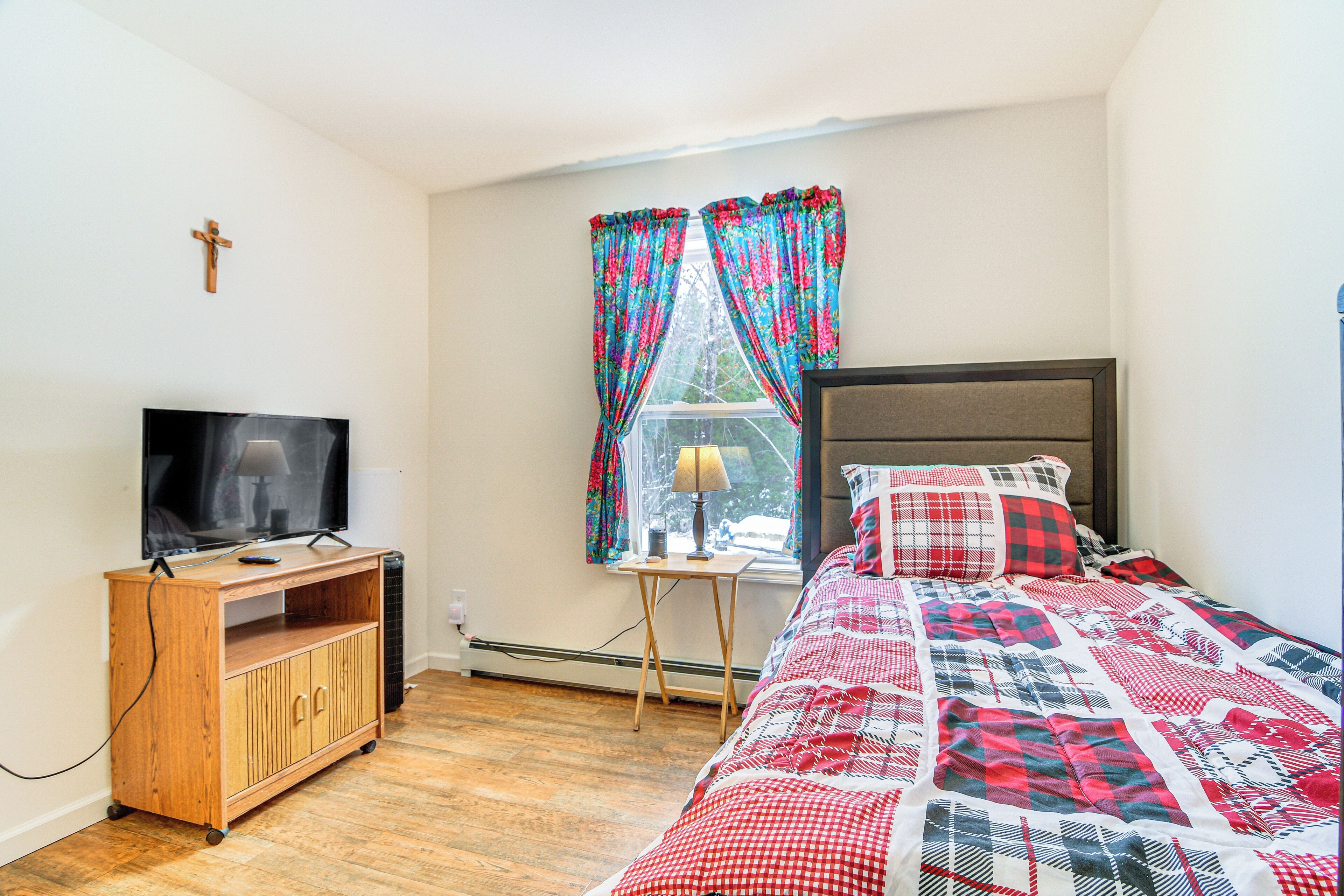 A bedroom with a bed, a TV on a wooden stand, a window with curtains, and a cross on the wall.