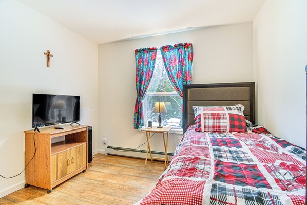 A bedroom with a bed, a TV on a wooden stand, a window with curtains, and a cross on the wall.