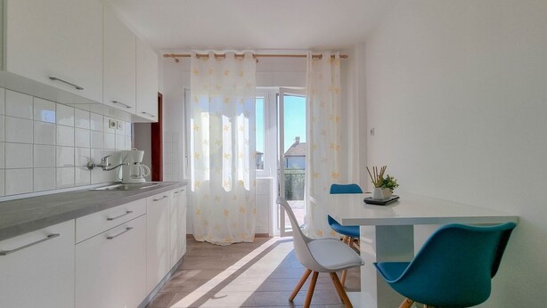 Una cocina moderna con armarios blancos, encimera gris y un comedor con una silla azul y una mesita.