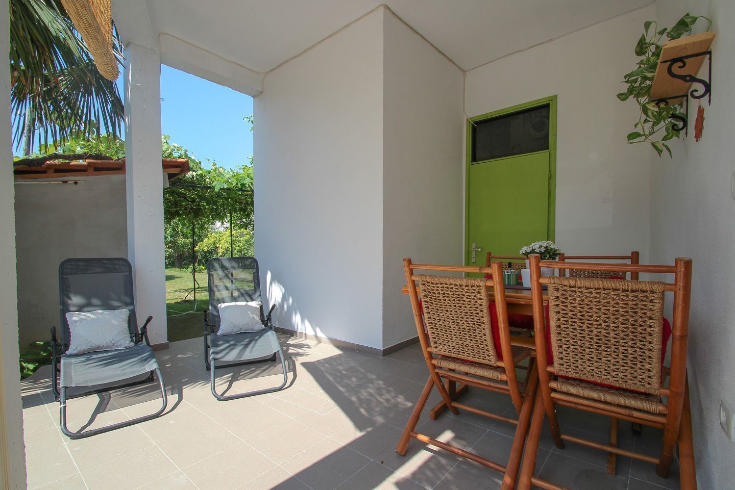 A covered patio area with two wicker chairs and a table, a green door, and a plant on the wall.