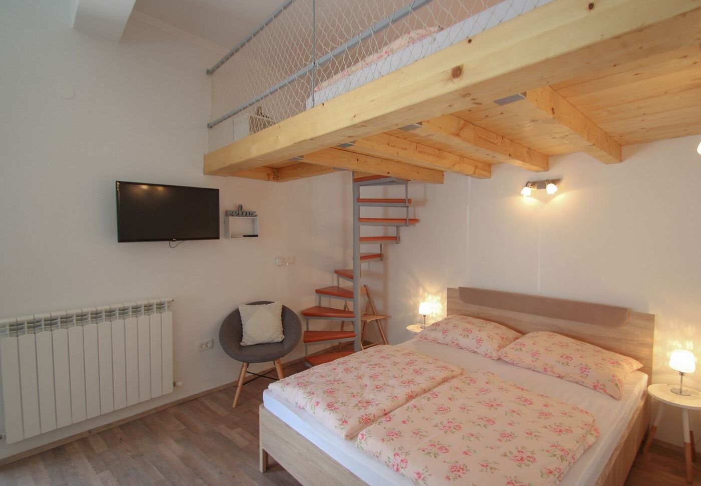 Ein Schlafzimmer mit Holzbalkendecke, einem Bett mit floralem Bettzeug, einem an der Wand befestigten Fernseher, einem Sessel, einem kleinen Tisch und einer Leiter zu einem Schlafpodest.