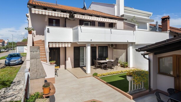 Ein zweistöckiges Haus mit Terrasse, einem geparkten Auto und einem kleinen Garten.