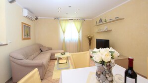 Ein Wohnzimmer mit einem beigefarbenen Sofa, einem kleinen Tisch mit Blumenvase, einem Fernseher und einer Weinflasche auf einem Tisch.