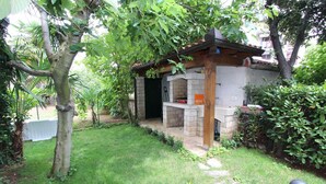 Un espace barbecue extérieur doté d’une structure en pierre, entouré d’une végétation luxuriante et d’une pelouse bien entretenue.