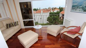 A living room with a sofa, ottoman, and chair, a small table, and a large sliding glass door leading to a balcony with a view of houses and trees.