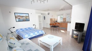 Un apartamento moderno y bien iluminado con sala de estar, comedor y cocina. Hay una cama con colcha estampada en azul y blanco, una mesa de centro blanca y un televisor sobre un mueble.