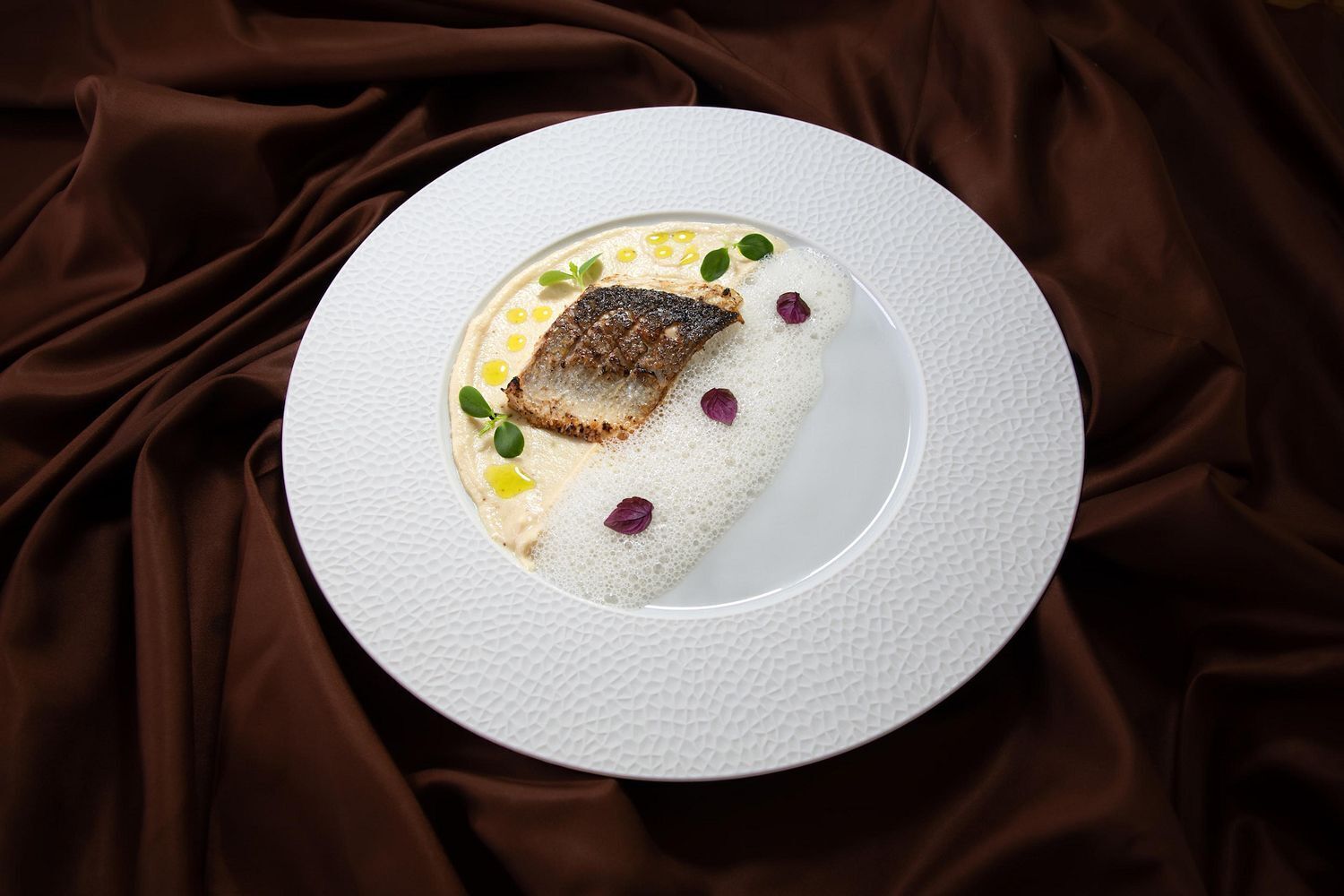 A white plate with a piece of fish, garnished with small leaves and edible flowers, on a textured white plate against a brown fabric background.
