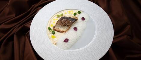 A white plate with a piece of fish, garnished with small leaves and edible flowers, on a textured white plate against a brown fabric background.