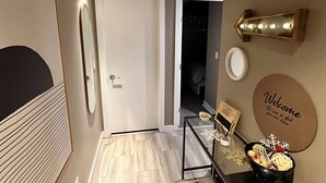 A hallway with a door, a mirror, a wall-mounted light fixture, a welcome sign, and a small table with a bowl of snacks. - Century park Condo Oasis (Edmonton)