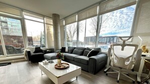 A modern living room with a grey sofa, a white coffee table, and a large window offering a view of buildings and trees. - Century park Condo Oasis (Edmonton)