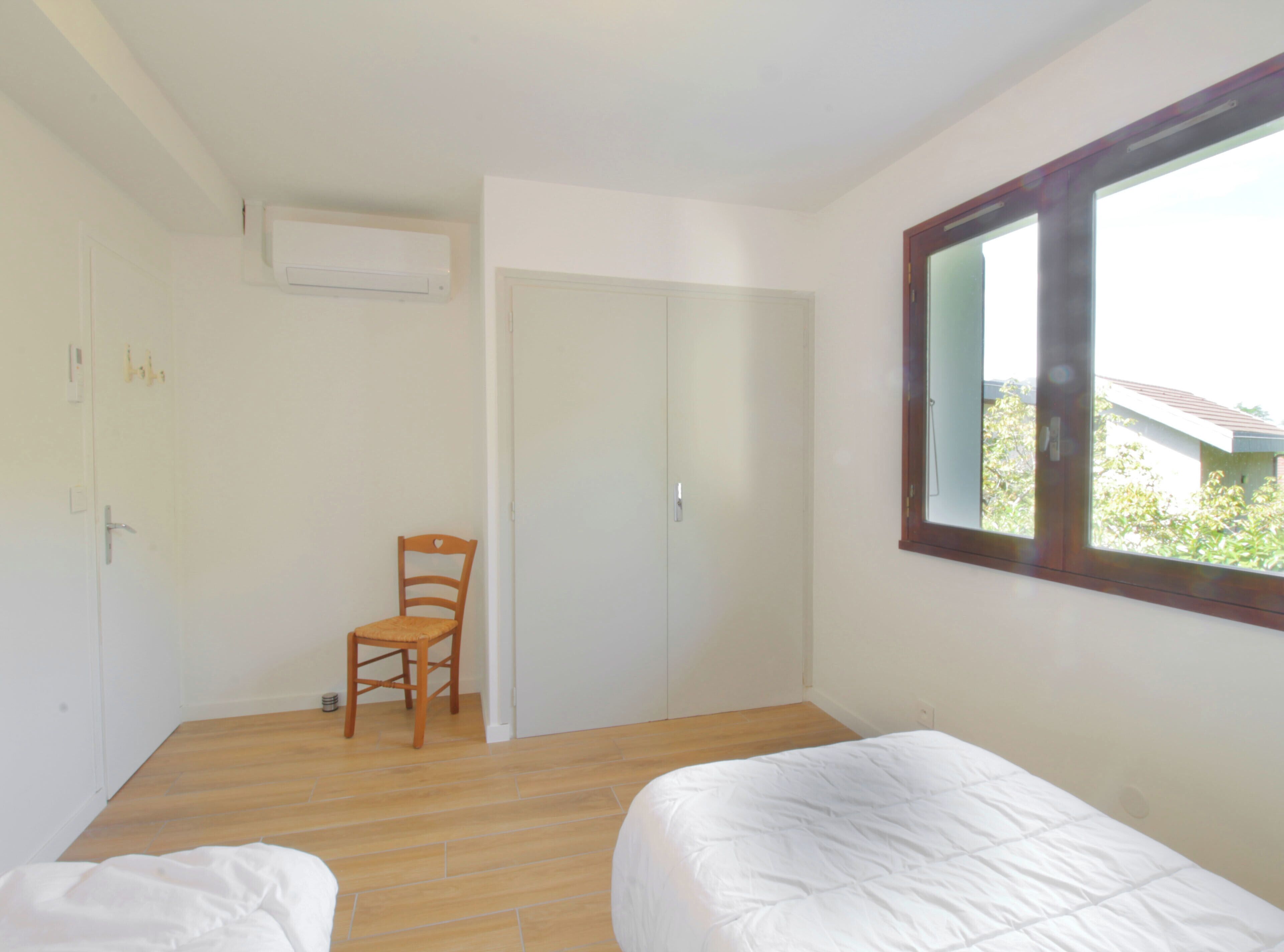 A bedroom with two beds, a wooden chair, a white wardrobe, and a window with a view of greenery outside.