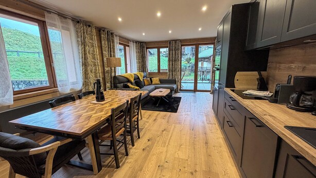 A modern kitchen with wooden flooring, a dining area with a wooden table and chairs, a sofa with yellow cushions, and a view of a green landscape through large windows. - Superb 3 room next to the chairlift in Arêches (Beaufort)