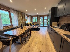 A modern kitchen with wooden flooring, a dining area with a wooden table and chairs, a sofa with yellow cushions, and a view of a green landscape through large windows. - Superb 3 room next to the chairlift in Arêches (Beaufort)