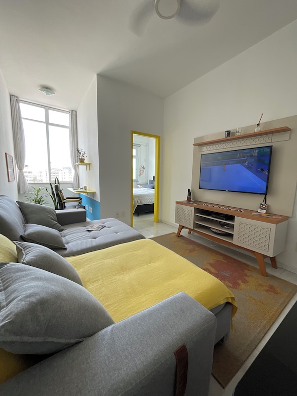 A modern living room with a large flat-screen TV, grey sofas, a yellow throw blanket, and a wooden entertainment unit.