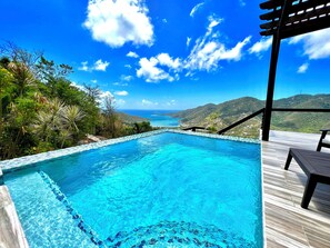Ein klares, blaues Schwimmbecken mit einer Holzterrasse und Liegestühlen, umgeben von üppiger Vegetation und einem malerischen Blick auf das Meer und die Hügel.