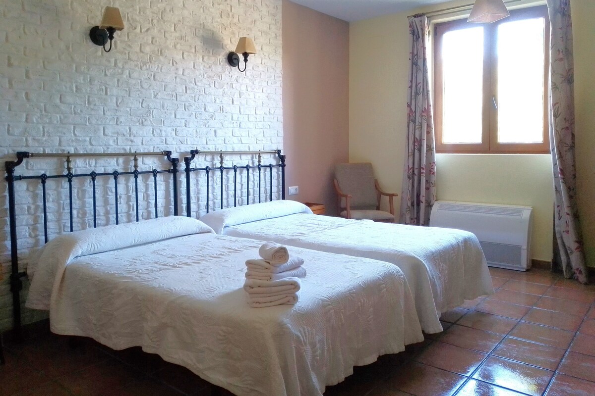 A room with two beds, a brick wall, a chair, a window with curtains, and a hanging light fixture.