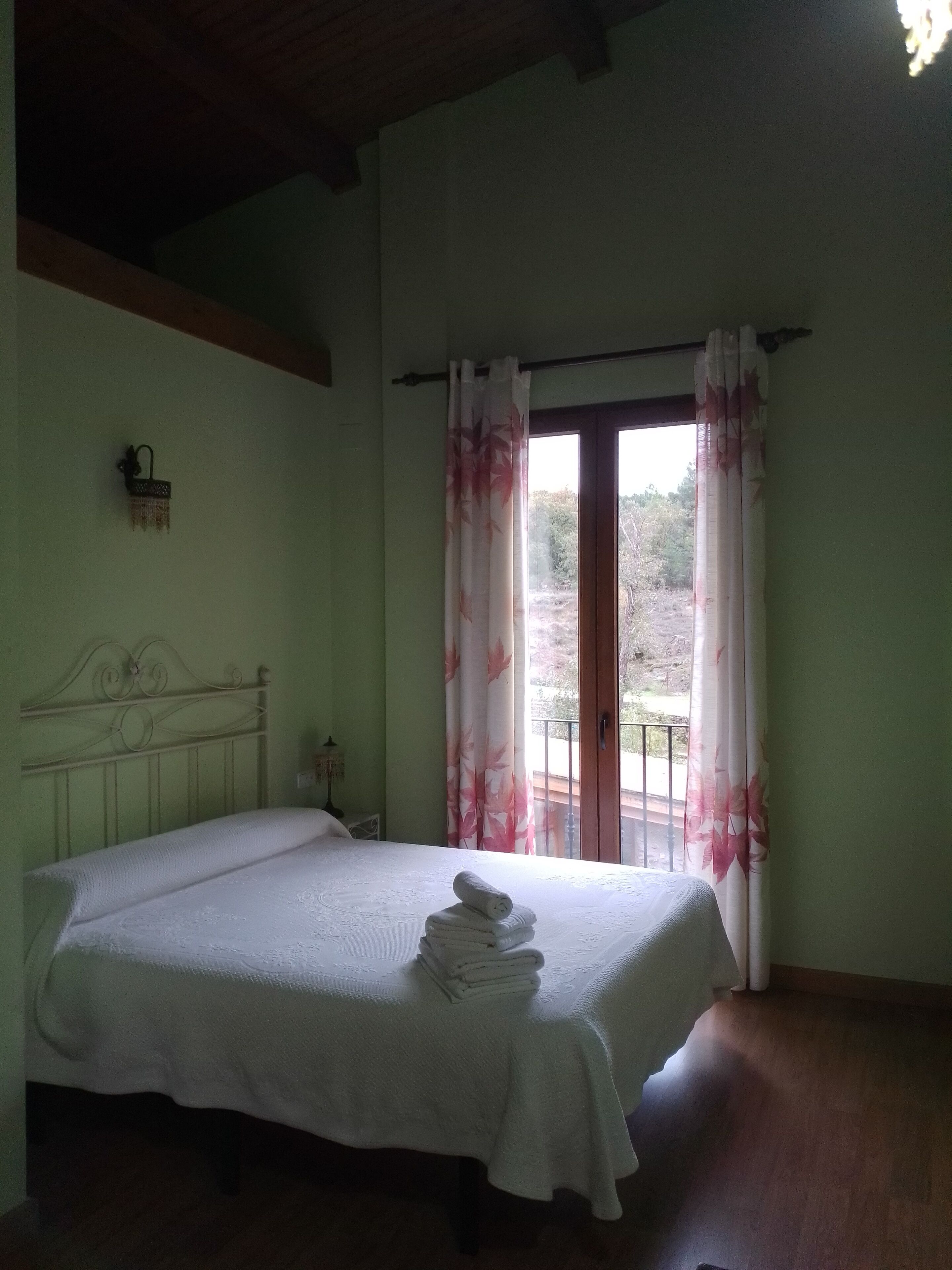 A bedroom with a bed, a nightstand, a wall-mounted lamp, and a window with curtains.