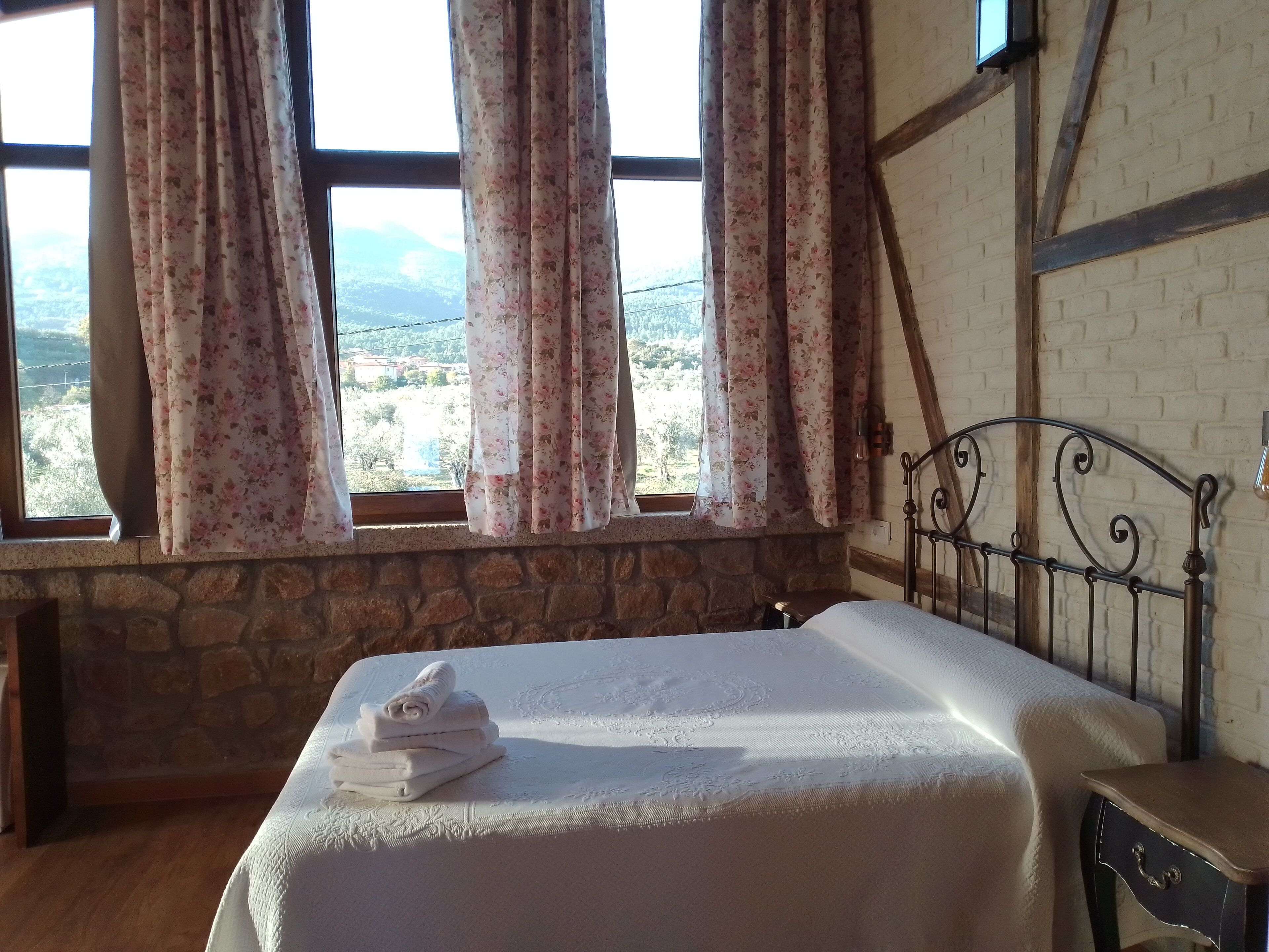Un dormitorio con pared de piedra, una cama con colcha blanca y vista a un paisaje montañoso a través de la ventana.