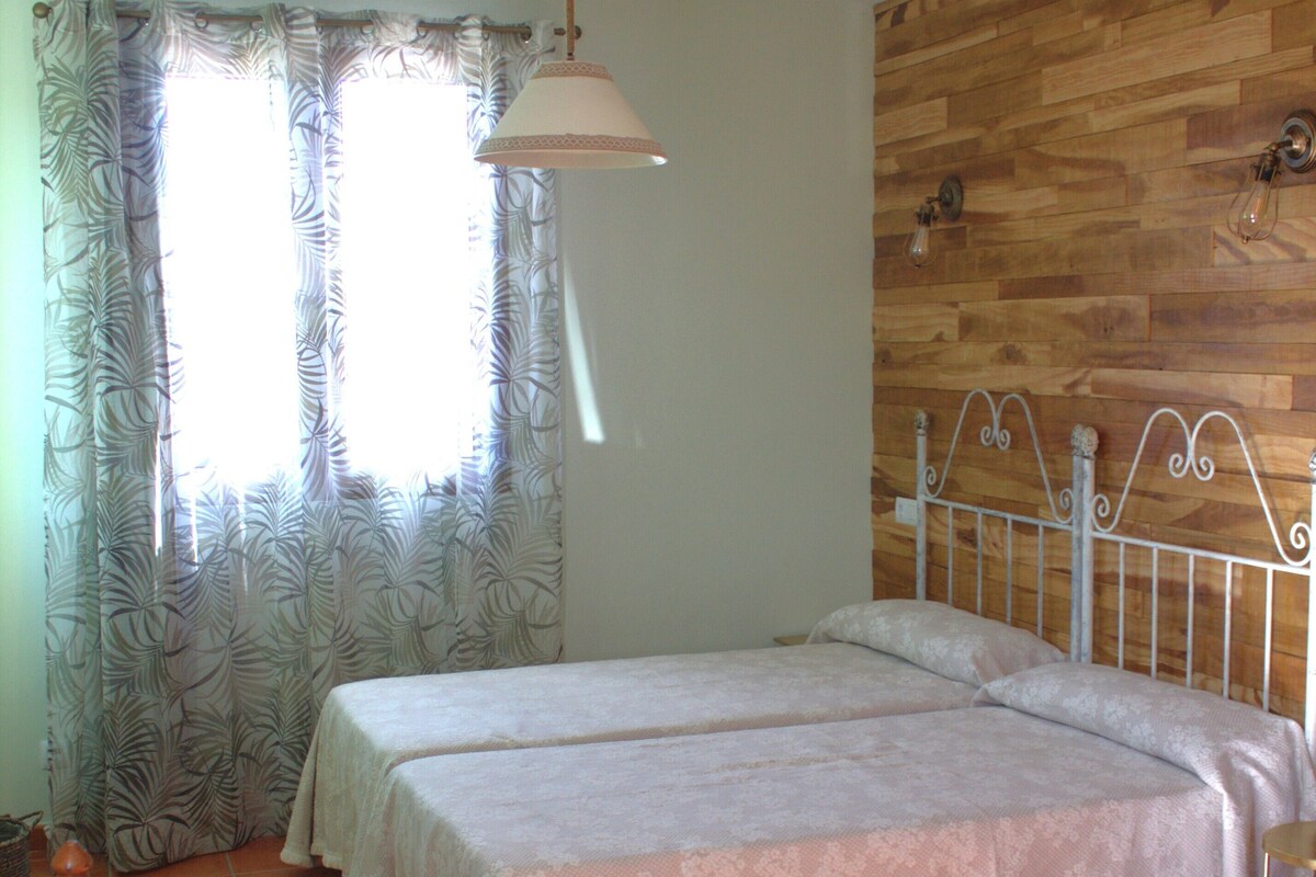 A bedroom with two beds, a wooden wall, a ceiling fan, and a window with curtains.