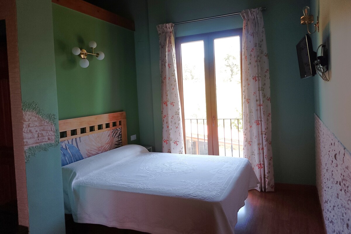 A bedroom with a bed, a window with curtains, a wall-mounted light fixture, and a wall-mounted electrical outlet.