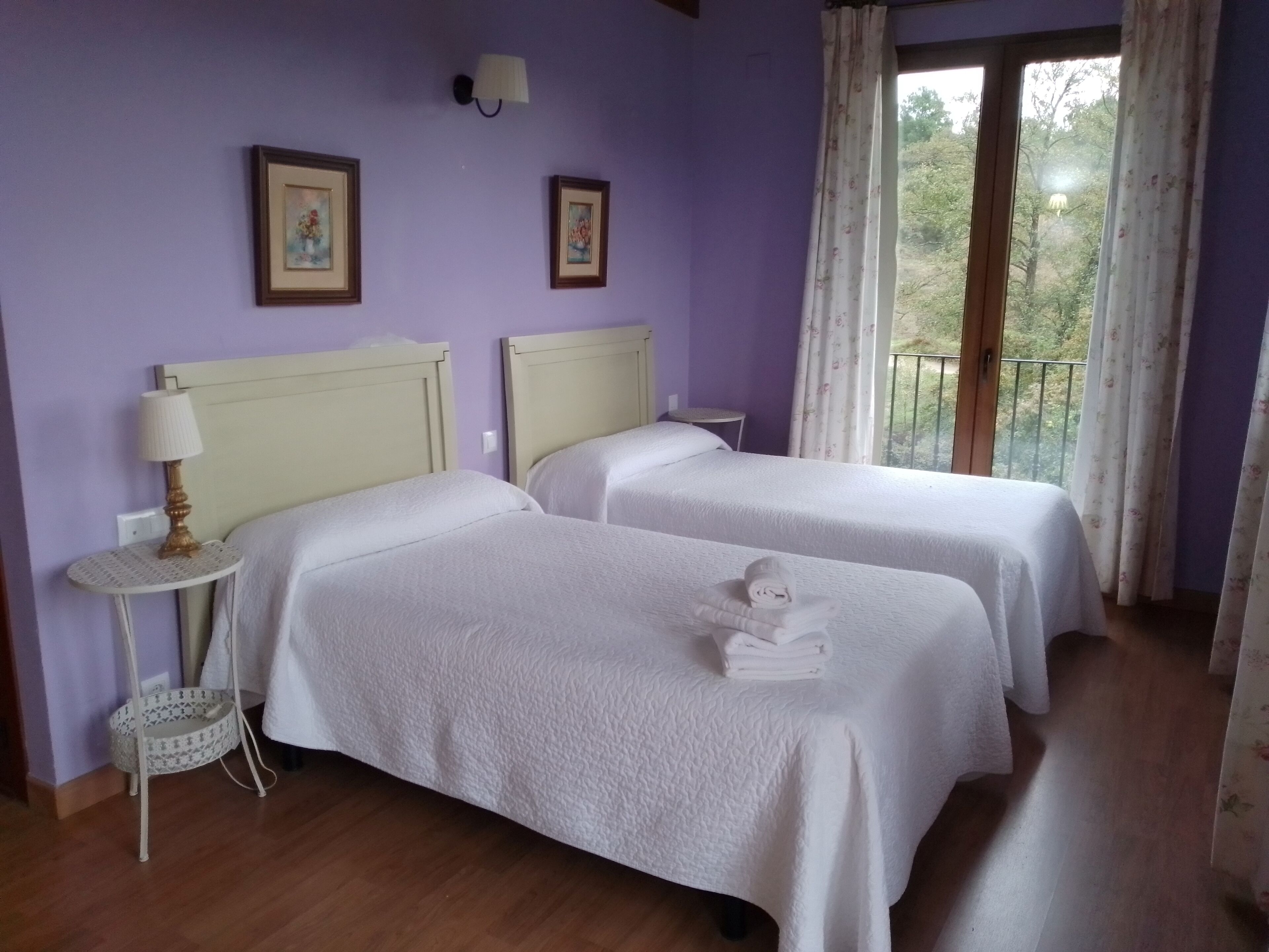 A room with two beds, a small table with a lamp, and a view of greenery outside.