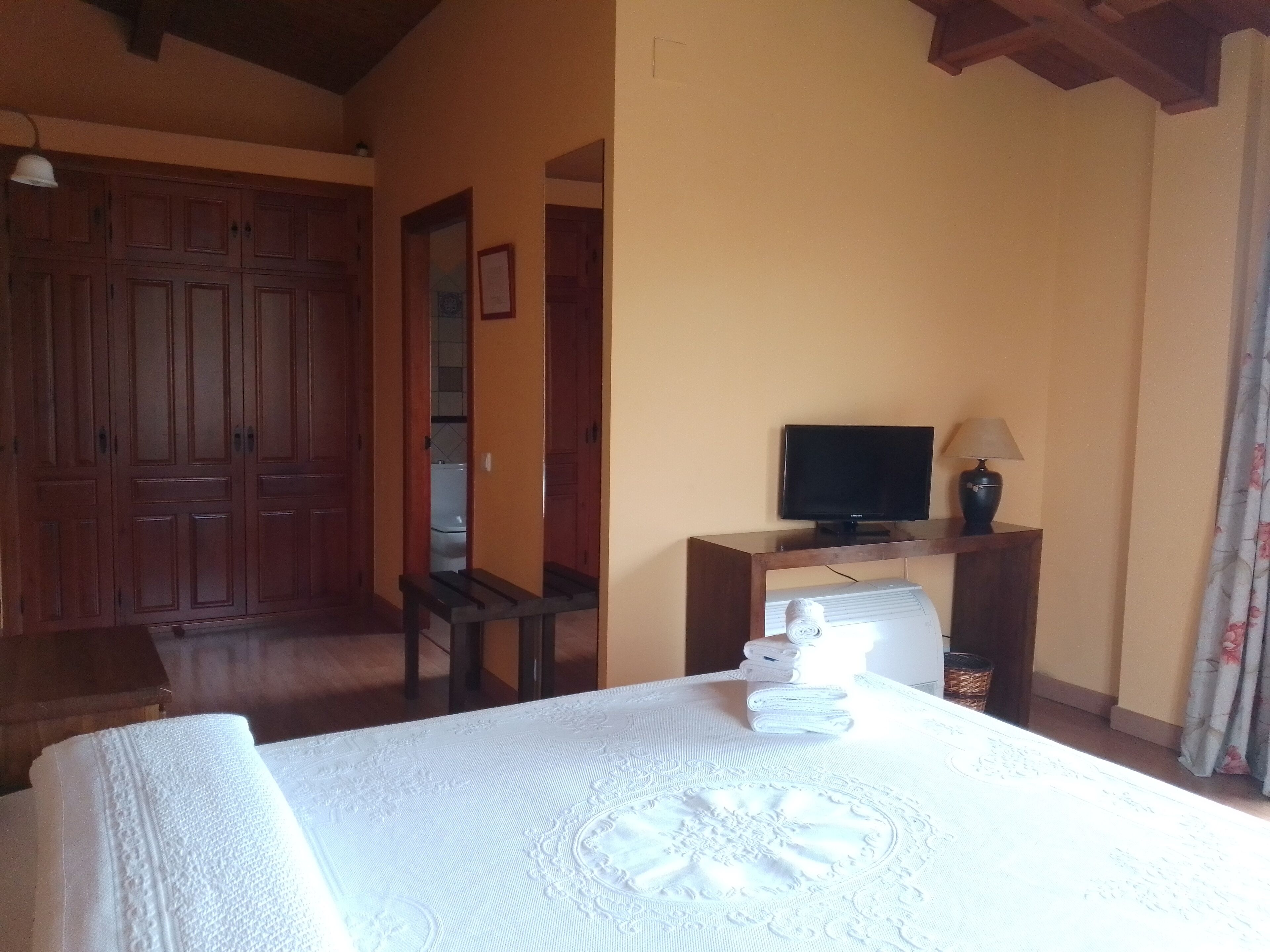A hotel room with a bed, a television on a wooden stand, a table with a lamp, and a wardrobe.