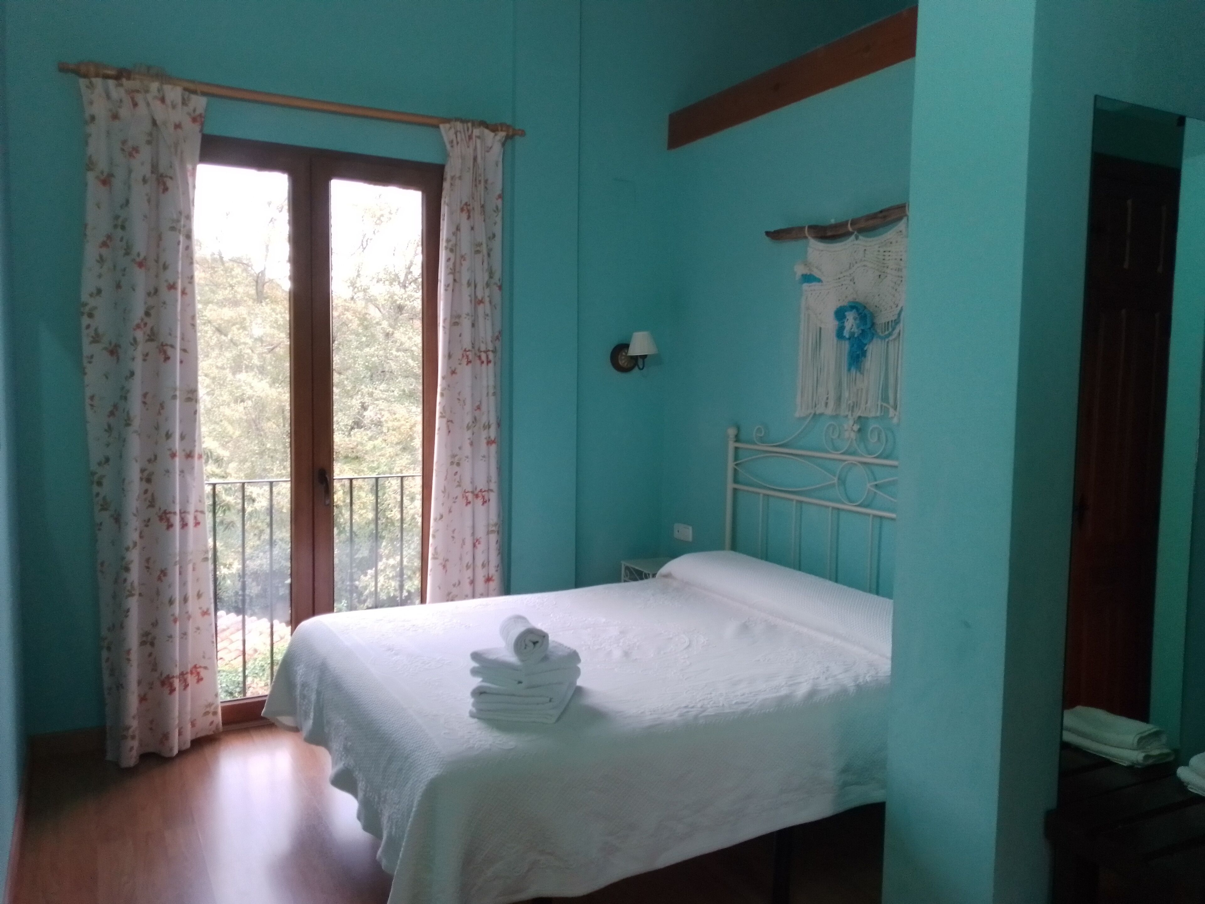 A bedroom with a bed, a window with curtains, a wall-mounted light, and a view of trees outside.