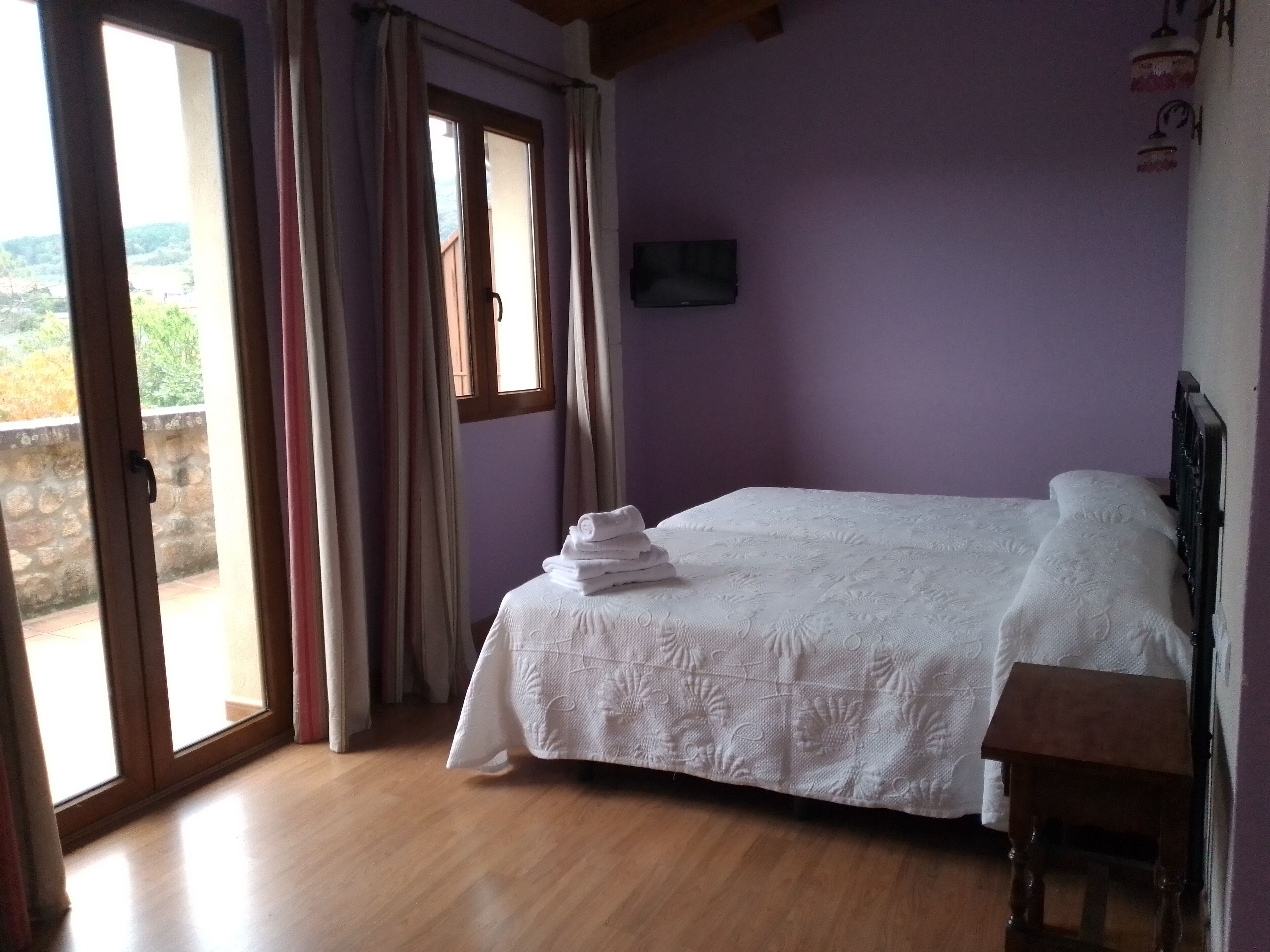 Un dormitorio con cama, mesita de noche de madera, televisor en la pared y vistas al exterior.