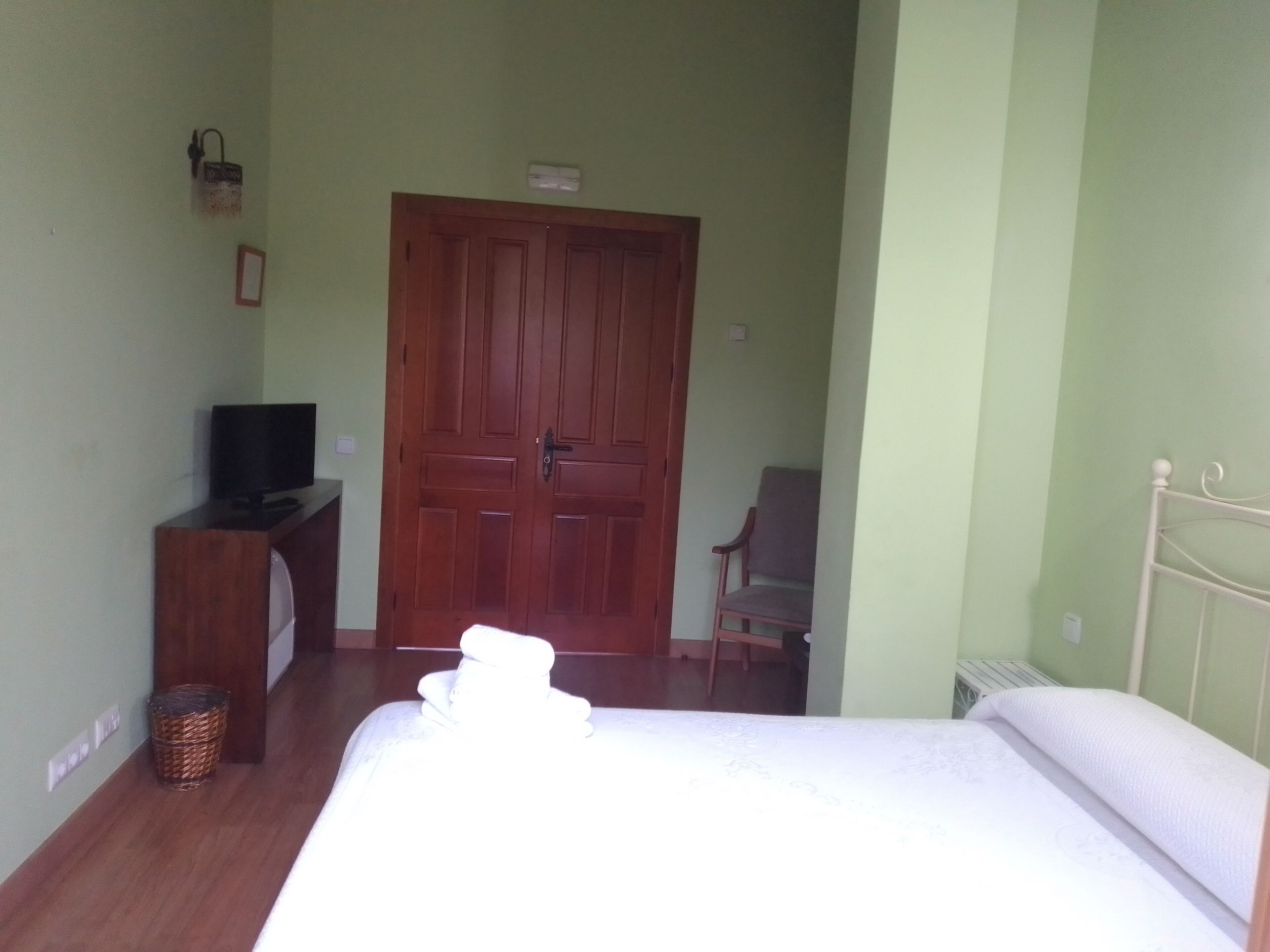 Une chambre avec une armoire en bois, un téléviseur posé sur un meuble en bois, un panier en osier, un lit blanc et une tête de lit en métal blanc.