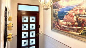 A hallway with a wooden door with geometric glass panels, a wall-mounted light fixture, and a framed painting of a coastal scene.