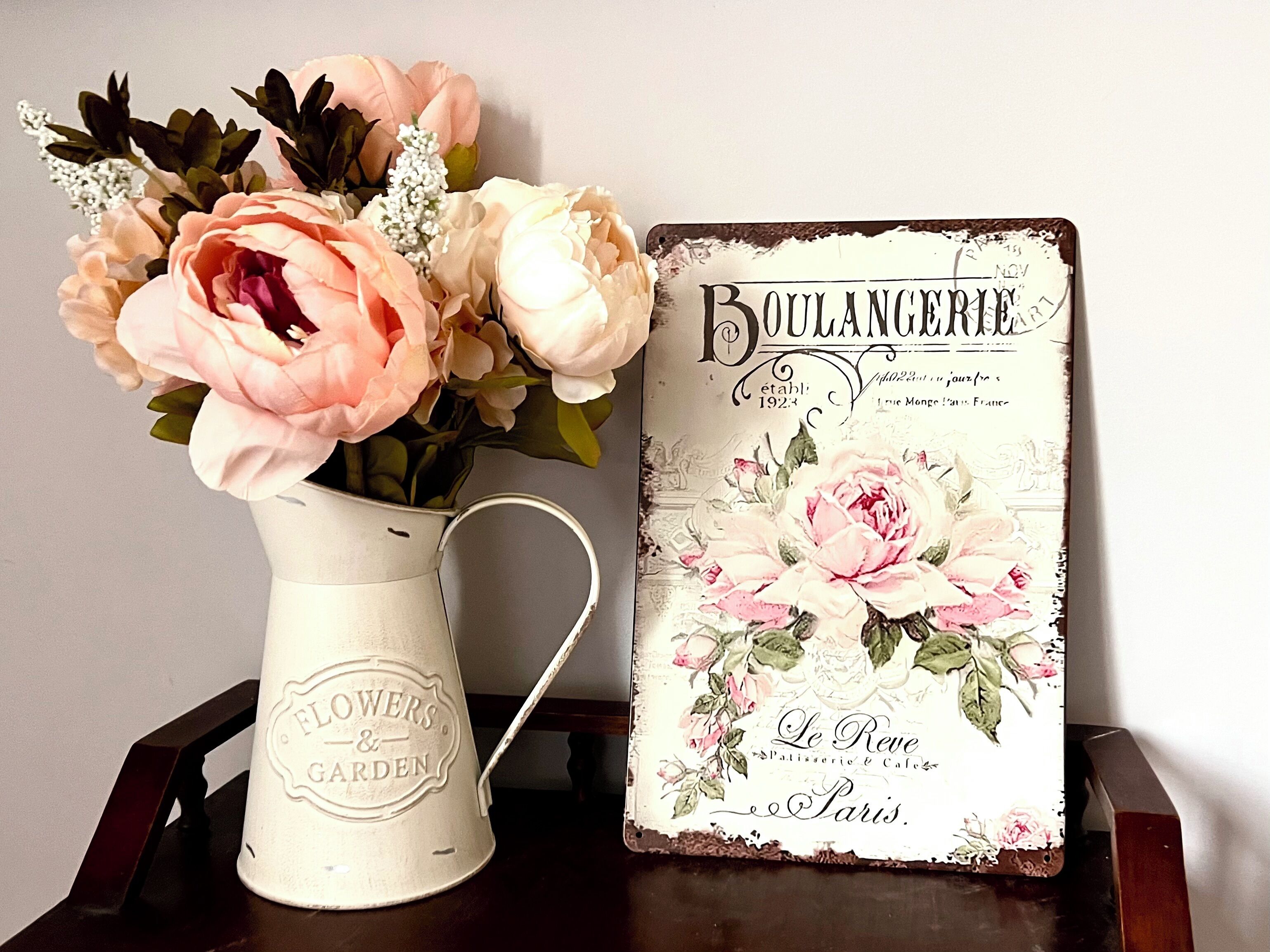 Ein Schild im Vintage-Stil mit floralen Elementen und Beschriftung, platziert neben einer weißen Metallvase mit Blumen.