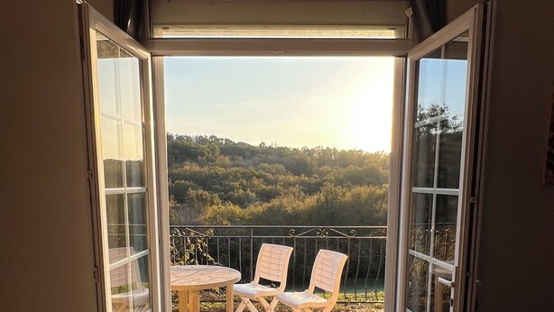 An open sliding door leading to a balcony with a table and two chairs, offering a view of a forested hillside.