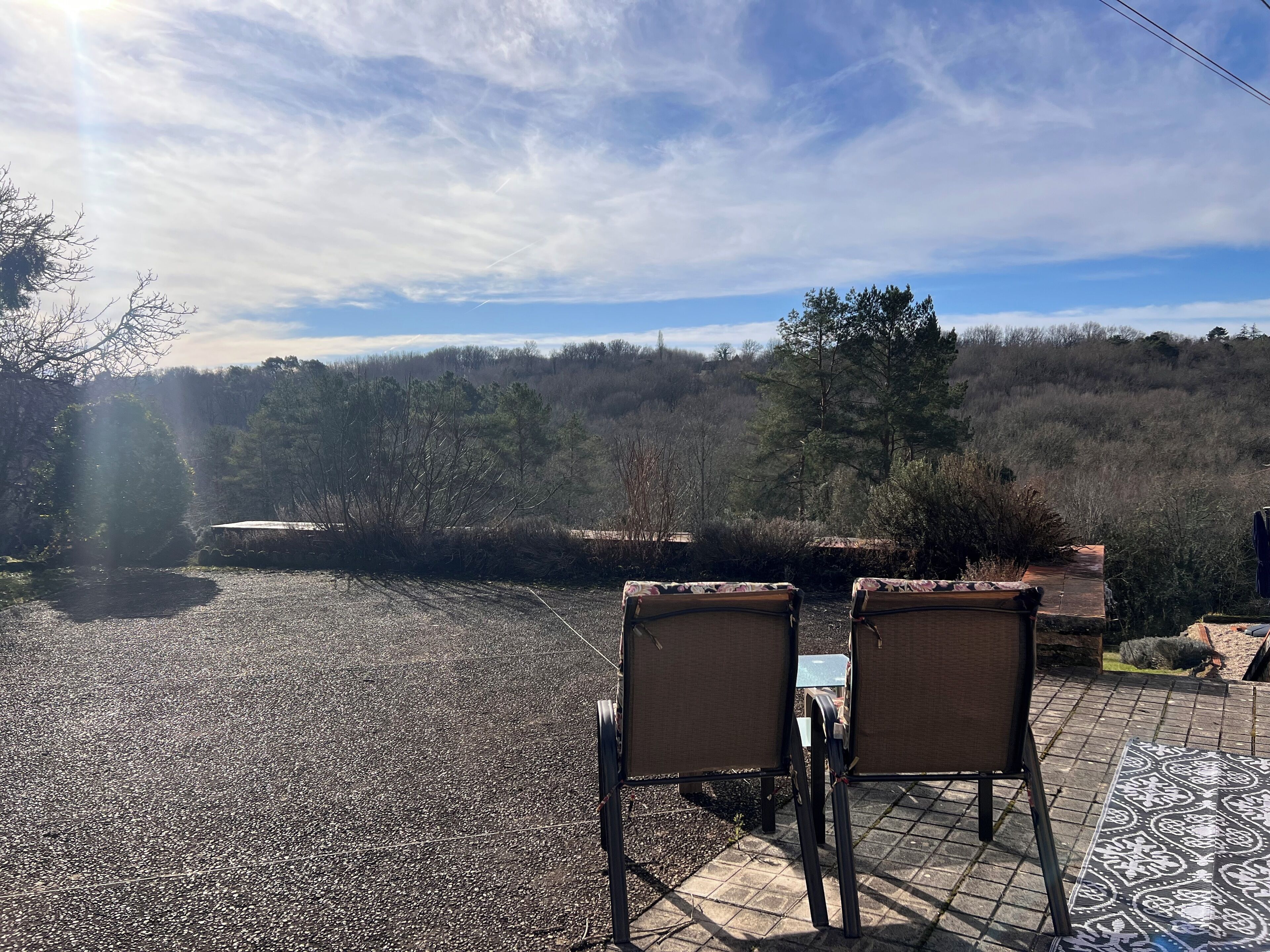 An outdoor seating area with two chairs and a table, overlooking a gravel path and a forested hillside.
