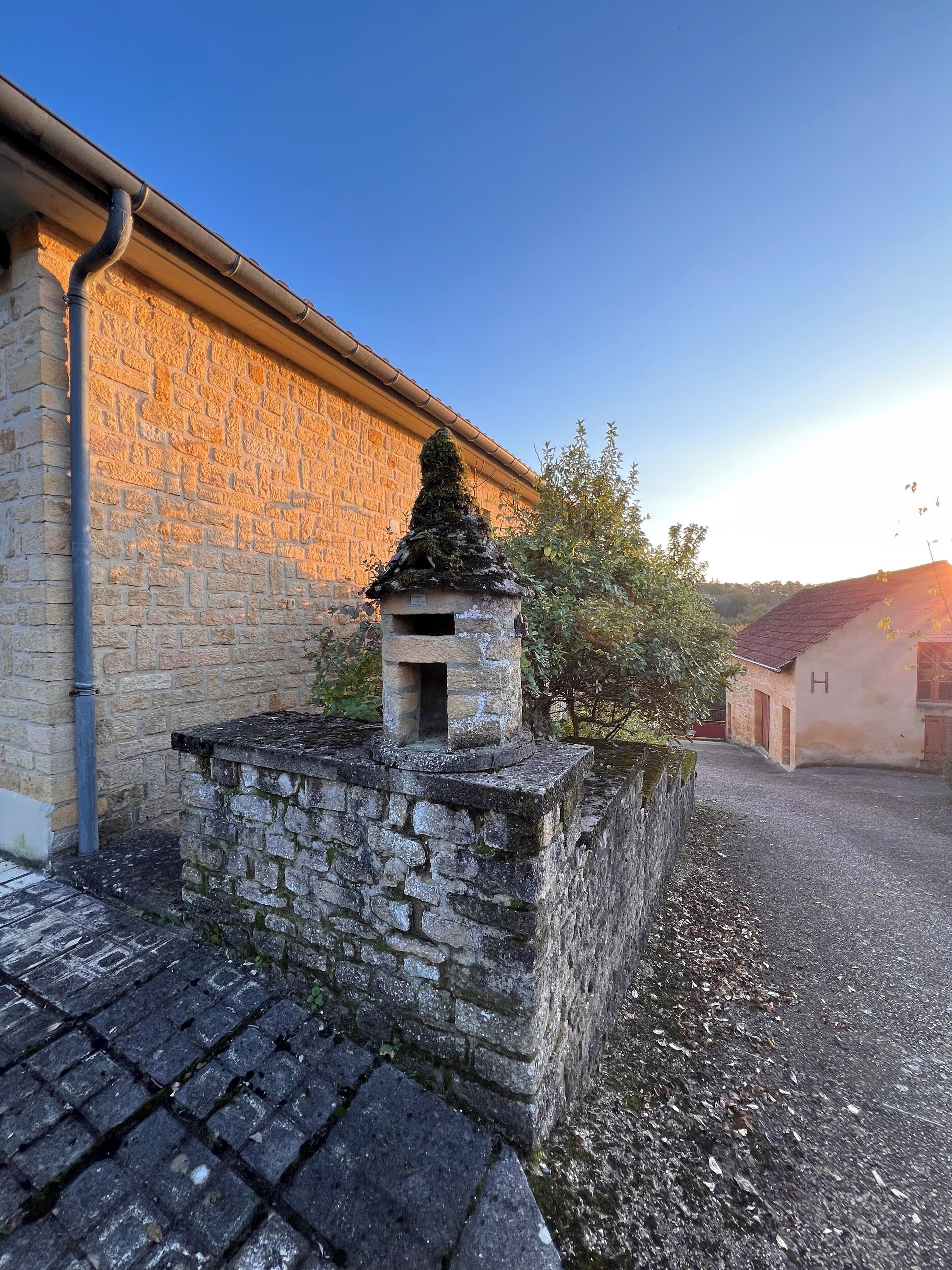 A stone wall with a small stone structure on top, a paved path, and a building with a red roof in the background.