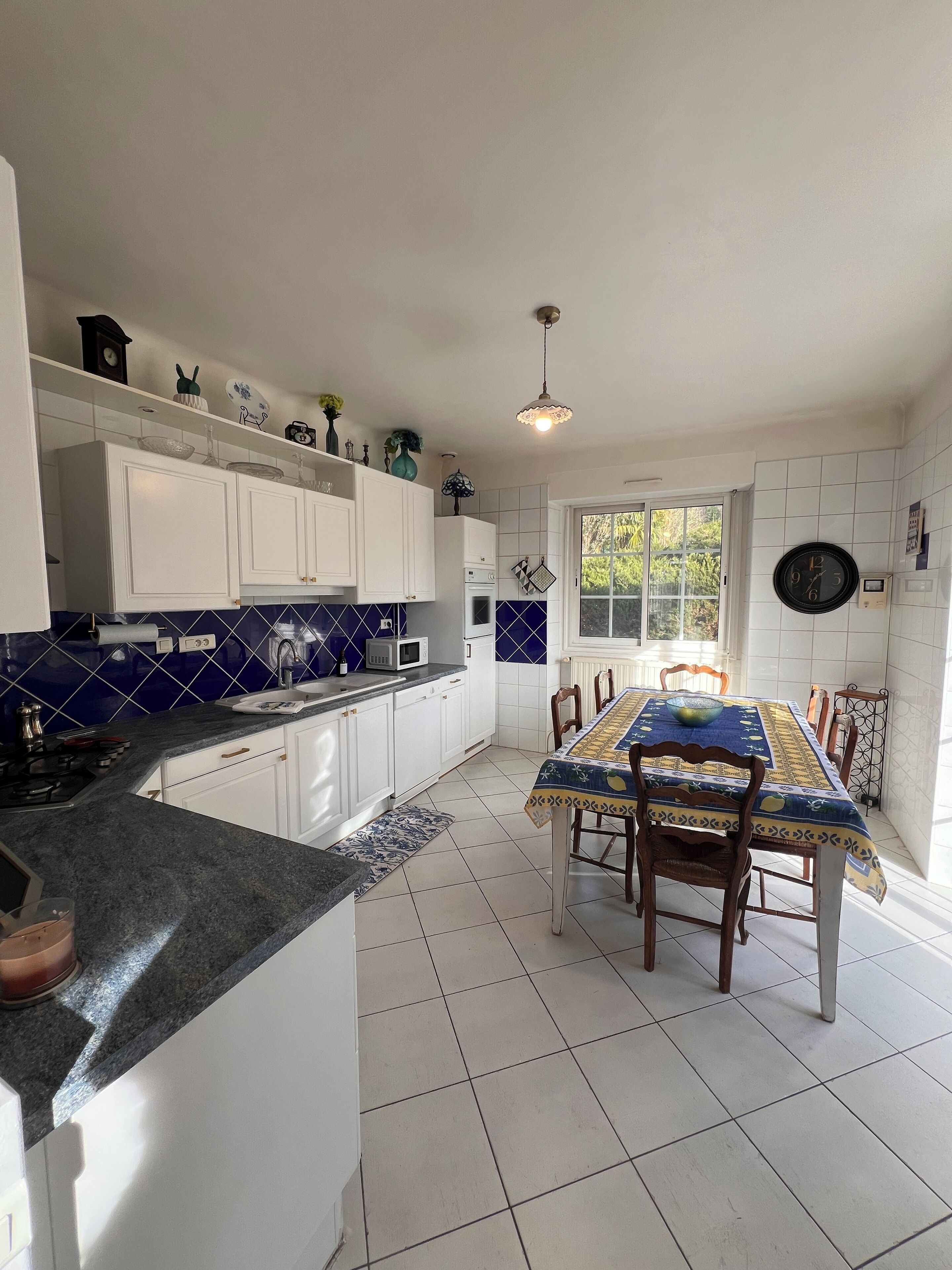 A kitchen with white cabinets, a blue tiled backsplash, a dining table with chairs, and a window with a view of greenery.