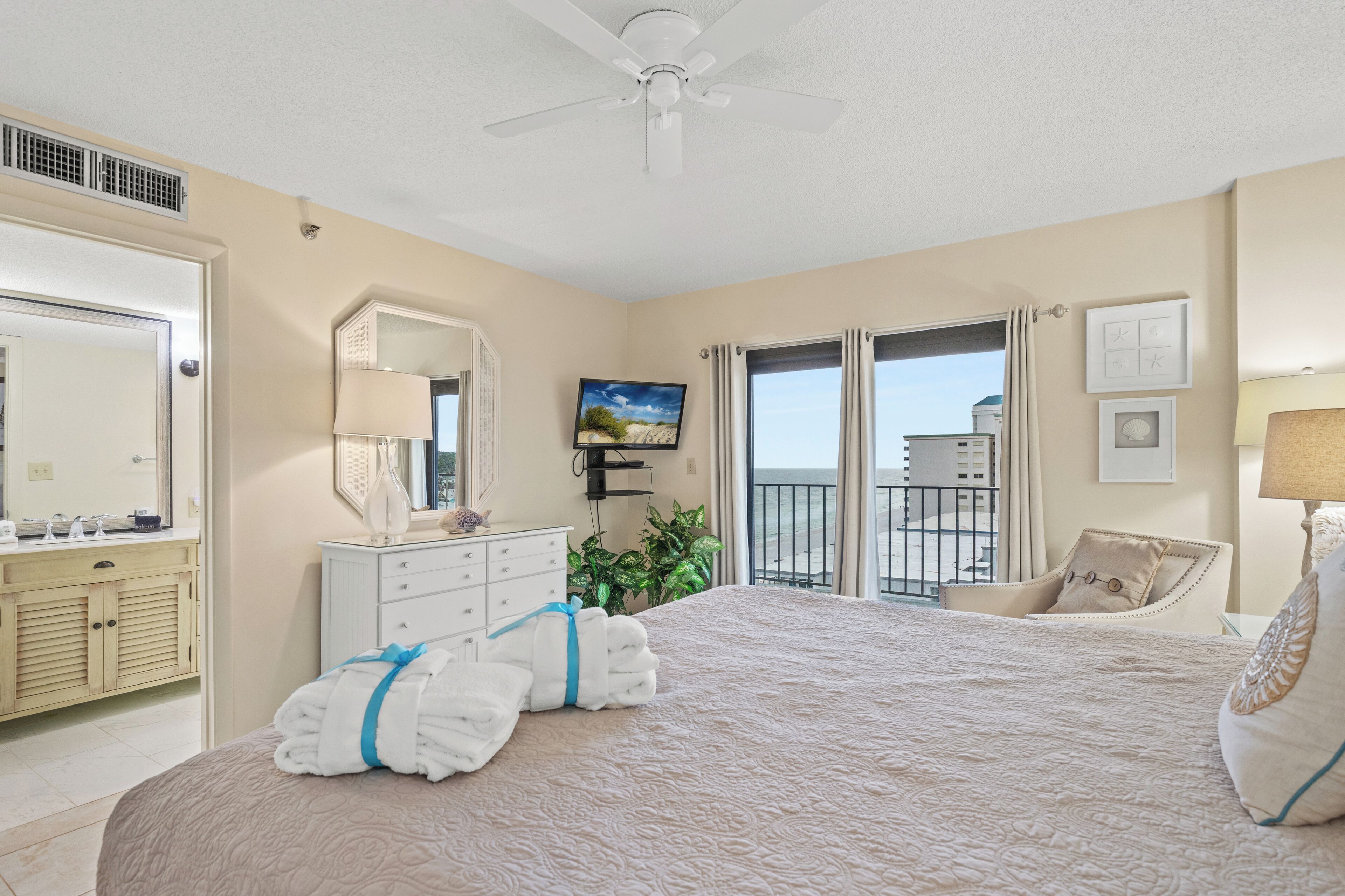 A hotel room with a large bed, a ceiling fan, a TV, a balcony with a view, and a bathroom with a vanity and mirror.