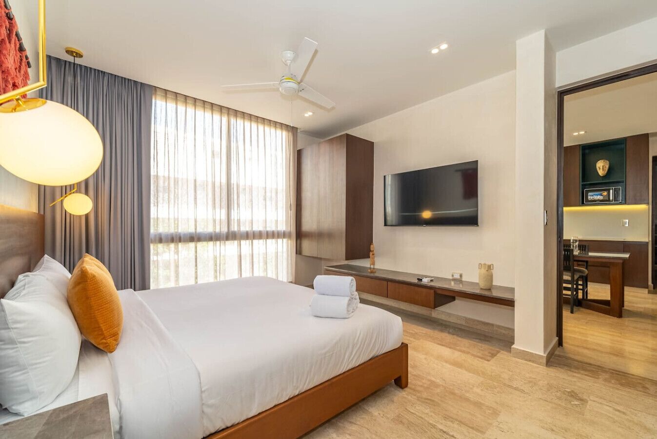 A modern hotel room with a large bed, a TV mounted on the wall, and a dining area visible through an open door.